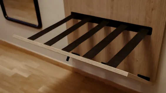 A folding luggage rack with black straps is extended from a wooden panel, set against a wall with hardwood flooring and a mirror nearby.