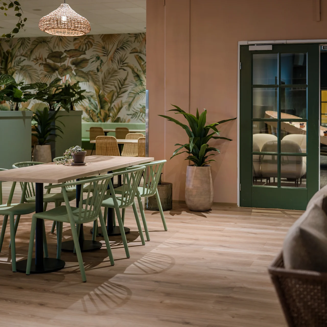 Wooden dining table with green chairs sits on a hardwood floor. Surrounding plants, rattan chairs, and tropical wallpaper contribute to a cozy indoor setting. A doorway leads to another room.