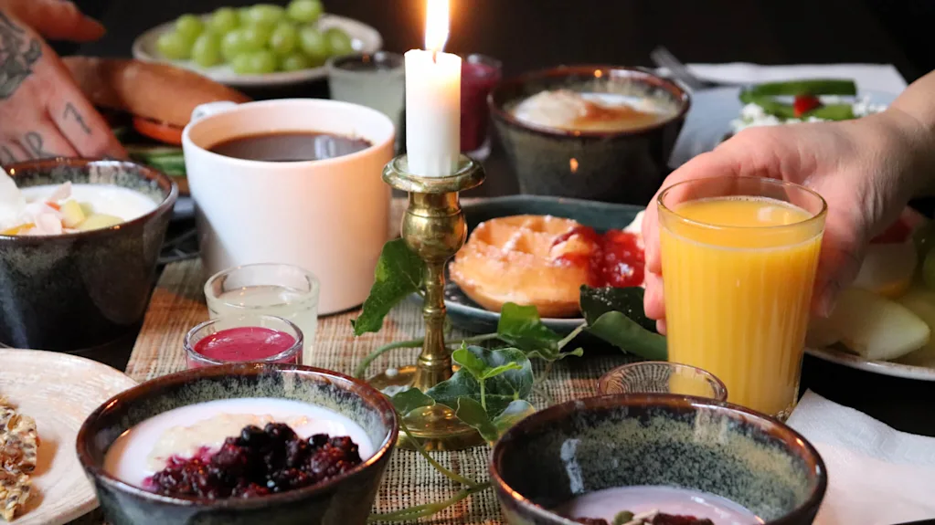 A breakfast table with various dishes, someone holding a glass of orange juice, lit candle, cozy atmosphere.