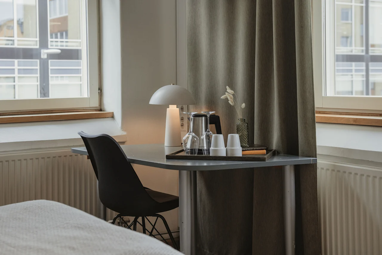 A well-lit desk area in a room at Home Hotel Uppsala, featuring a lamp, glasses, and a comfortable chair by a window.
