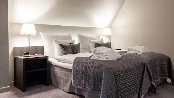 Two neatly made beds covered with grey quilts, flanked by lamps on bedside tables, in a softly lit room with slanted ceiling.