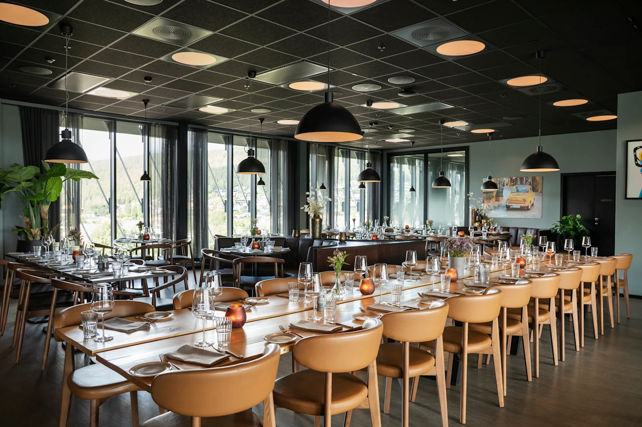 Et moderne restaurantområde med lange bord dekket for gjester, med store vinduer som vender ut mot en naturskjønn utsikt.