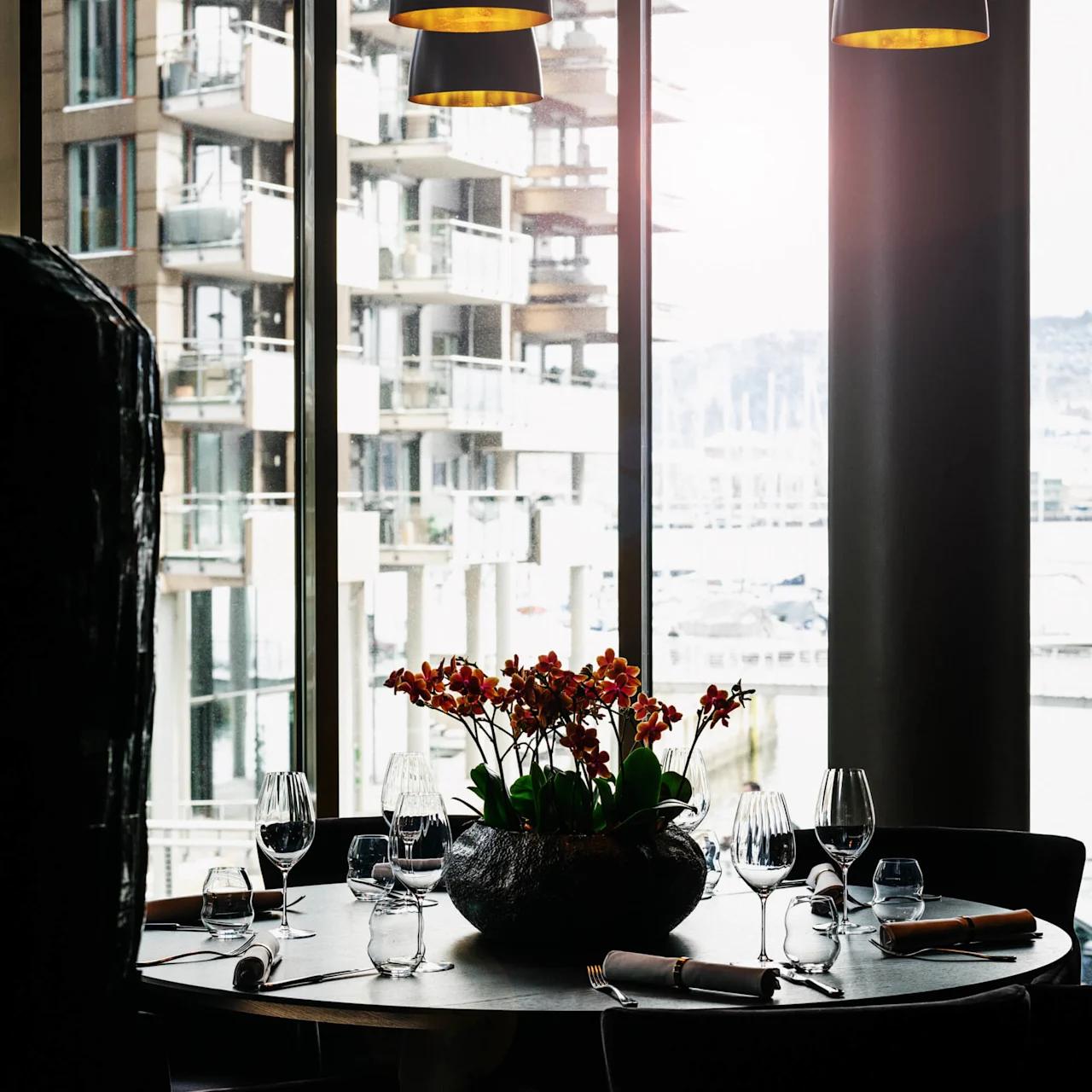 Et rundt bord dekket for middag med glass, bestikk og en dekorativ plante står ved store vinduer med utsikt mot moderne leilighetsbygg.