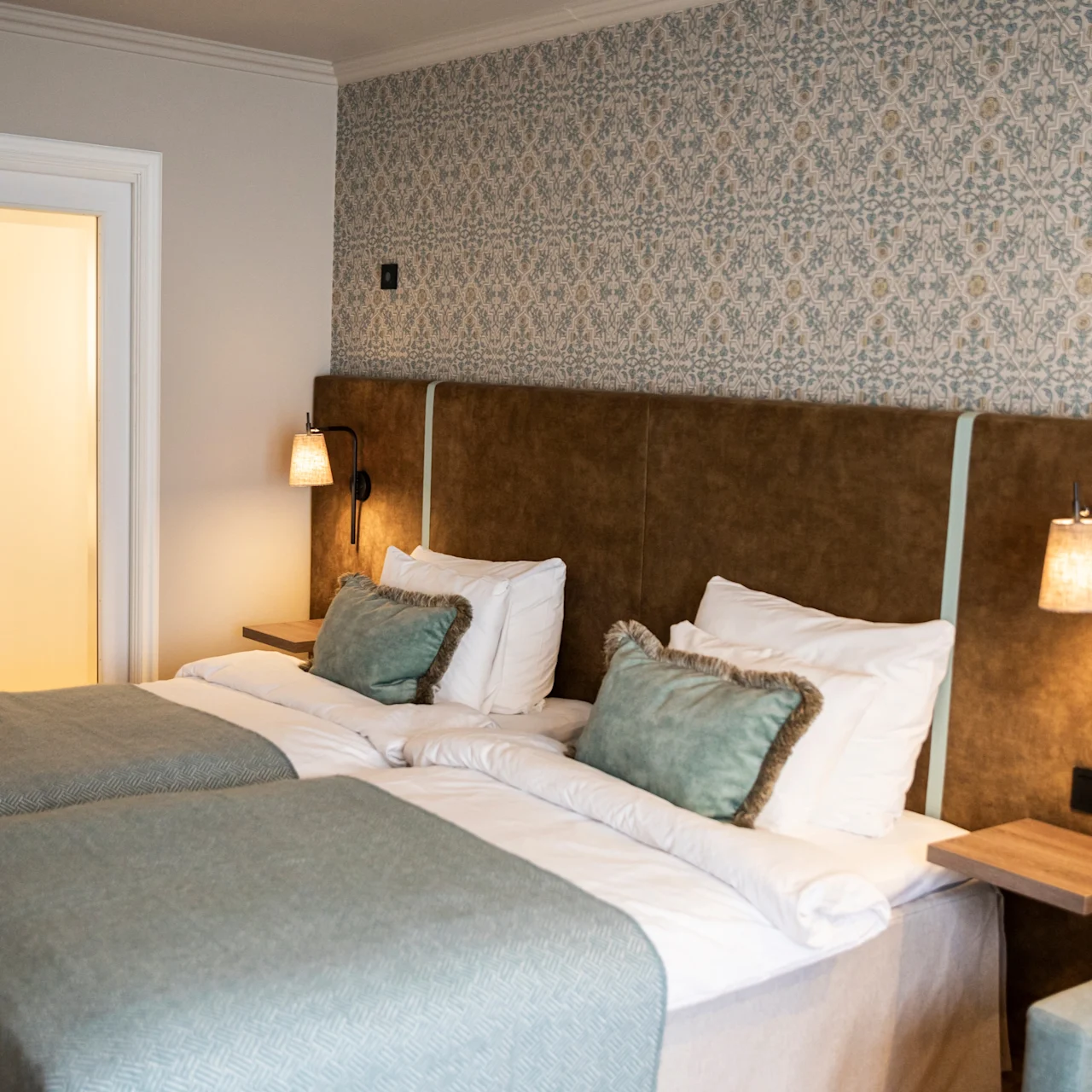 Two neatly made twin beds with light blue covers and green cushions are situated in a hotel room, featuring patterned wallpaper and individual bedside lamps mounted on the padded headboards.