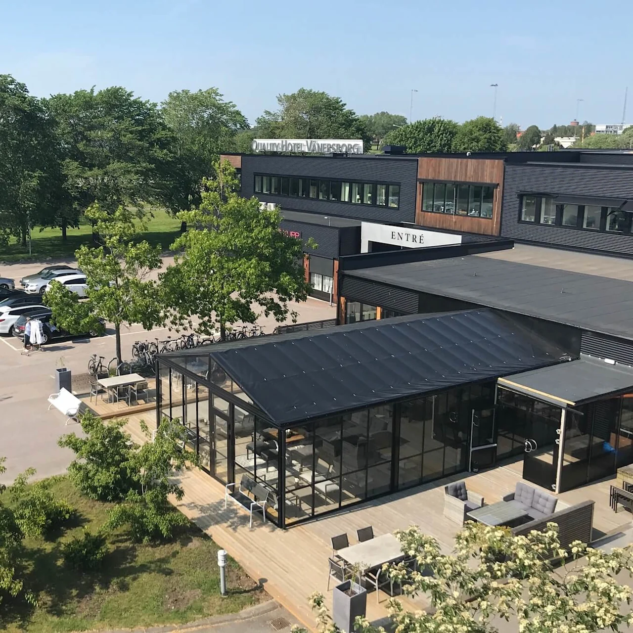En moderne bygning mærket "Quality Hotel Vänersborg" med en glasudvidelse står ved siden af en parkeringsplads fuld af biler. Omgivende træer og plæner forbedrer det urbane miljø.