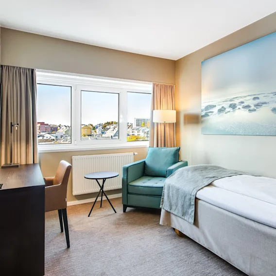 Lyst hotellrom med enkeltseng, blågrønn lenestol, skrivebord, havkunst på veggen og utsikt over Kristiansund.