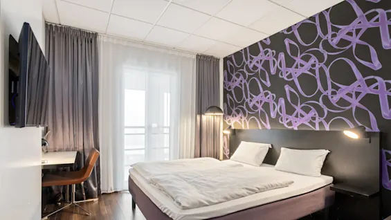 A double bed dominates a modern hotel room, accented with a purple headboard design, white bedding, and wooden flooring.