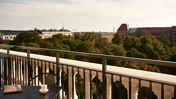 Et bord med en kopp og en mappe står på en balkong med utsikt over frodige grønne trær og fjerne klassiske bygninger under en klar himmel.