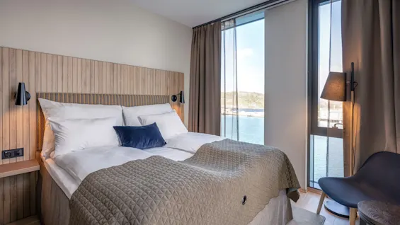 En seng med hvitt sengetøy og et grått teppe, i et lyst rom med treverk, har utsikt over rolig vann gjennom store vinduer.