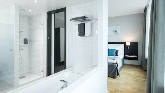 Et moderne bad med glassvegg dusj og badekar, koblet til et soverom med en pent oppredd seng, tregulv og et nattbord med lampe.