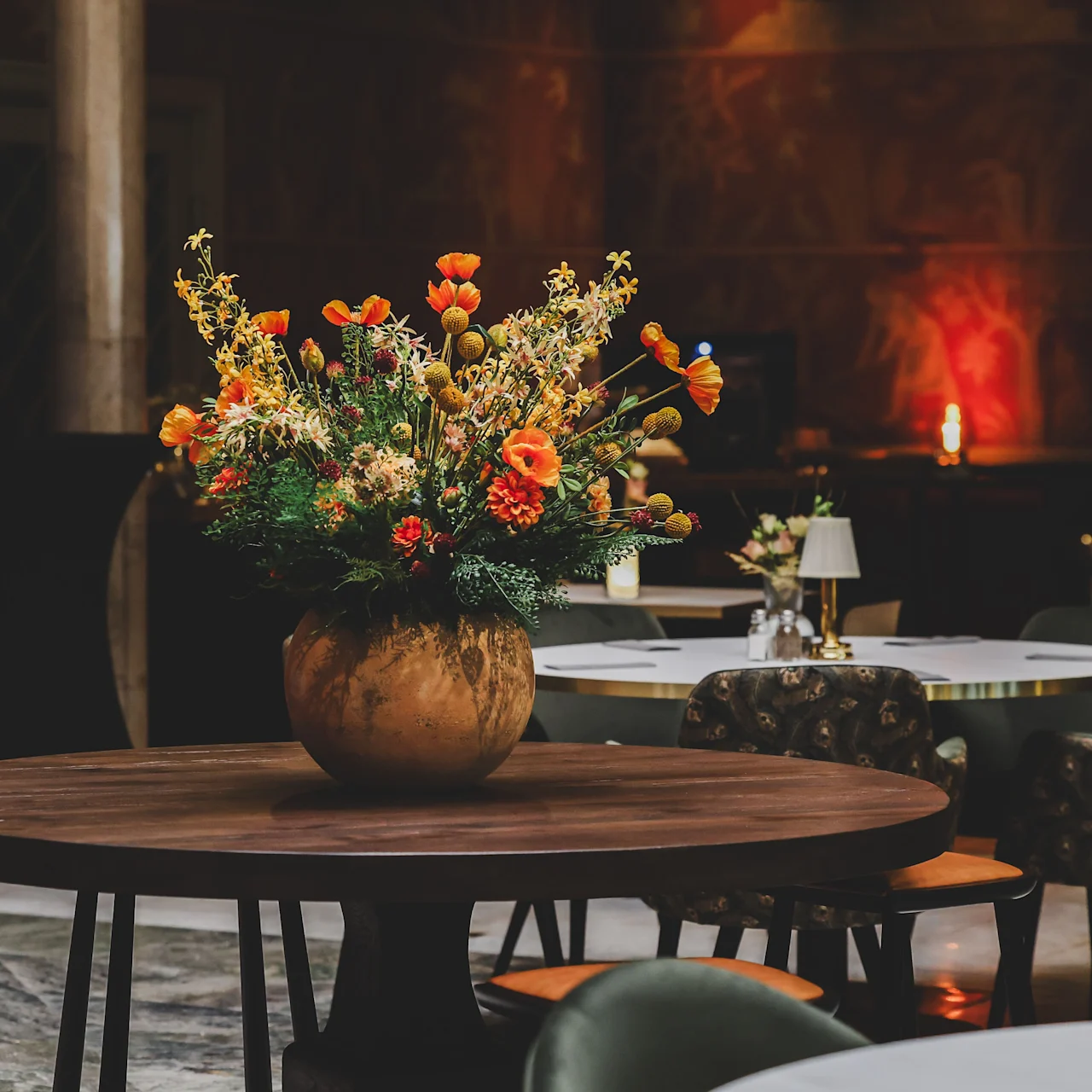 A large bouquet of vibrant flowers sits in a round vase on a wooden table, surrounded by cushioned chairs in a softly lit, elegant dining area.