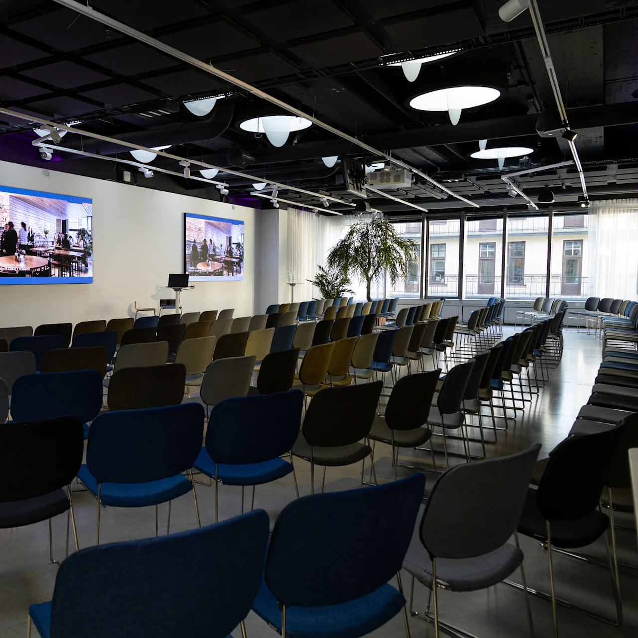 Rows of empty chairs face two large screens on a wall in a brightly lit conference room, decorated with potted plants and windows showing an exterior building view.