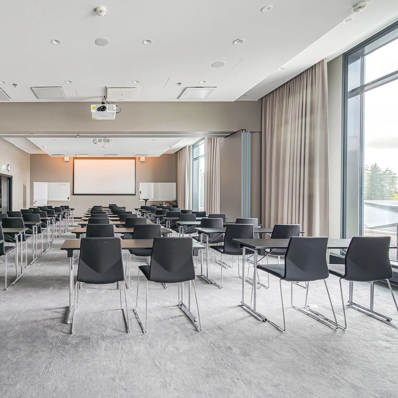 Rows of black chairs and tables face a large projection screen in a well-lit, modern conference room. Large windows and curtains line the right side.