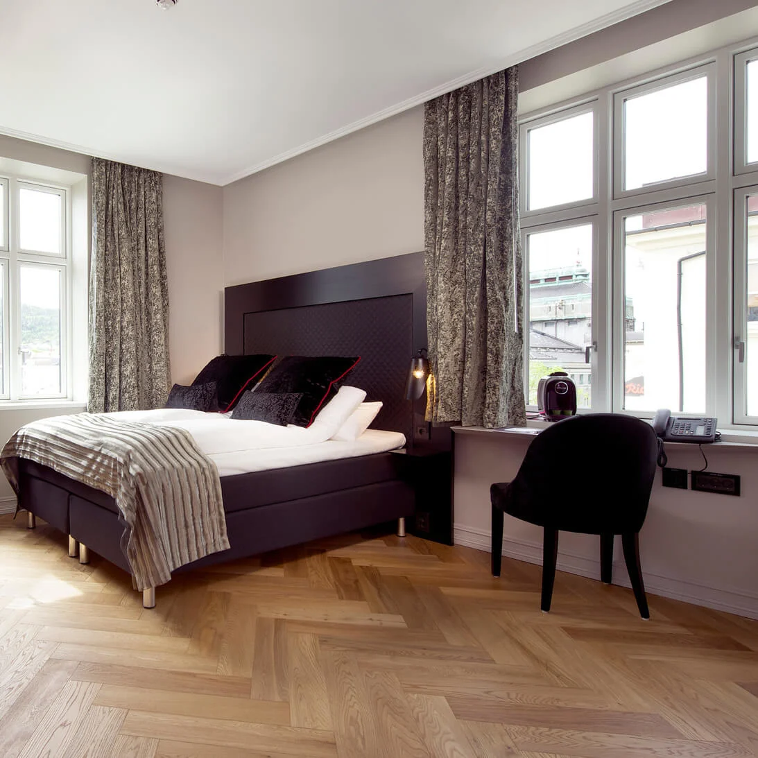 A bed with striped blanket and cushions stands in a modern room with large windows, wooden floors, and velvet chairs. A phone and coffee machine are on the windowsill.