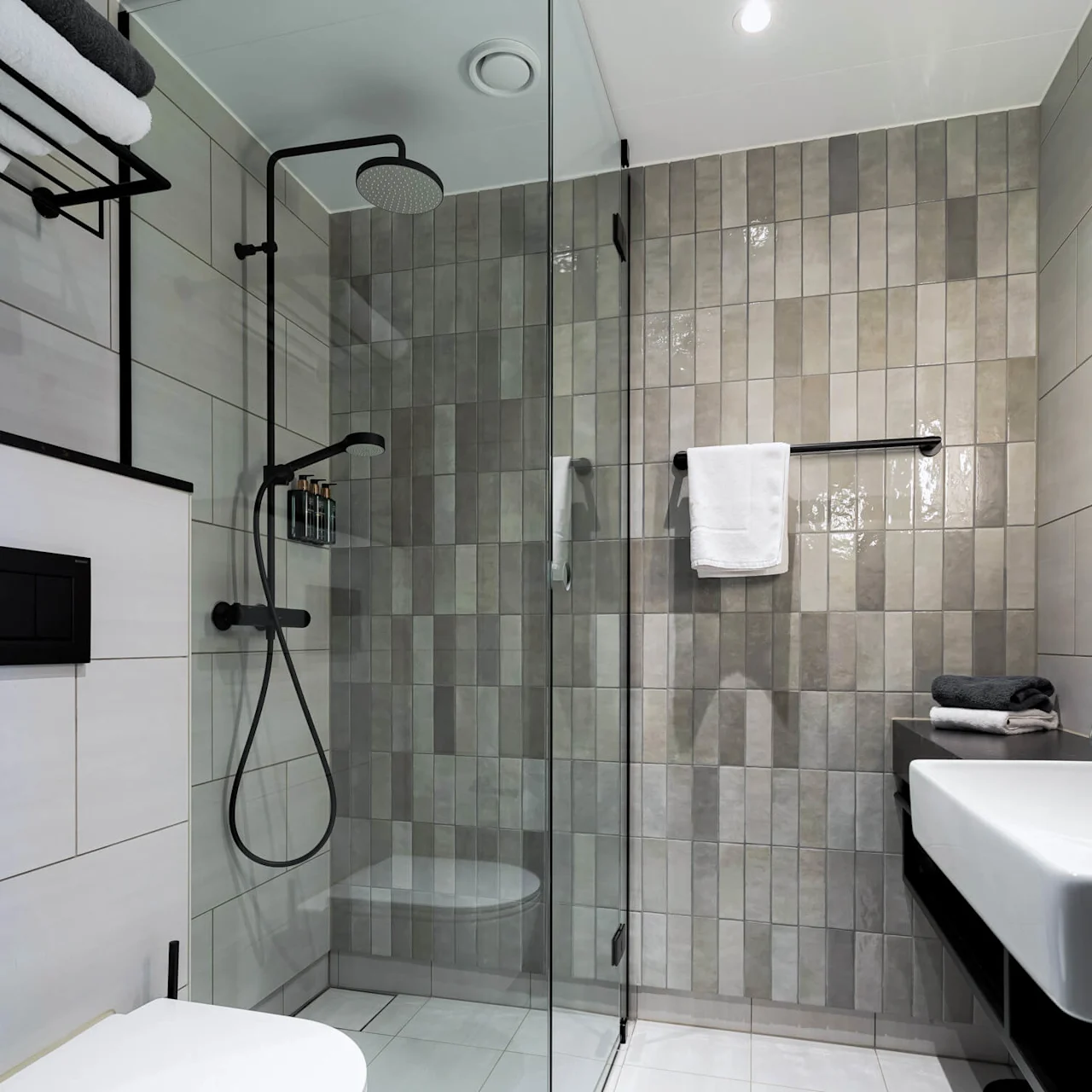 A modern bathroom features a glass shower with black fixtures. Towels rest on a rack and a sink is adjacent. Beige tiles cover the walls, providing a sleek, minimalist look.