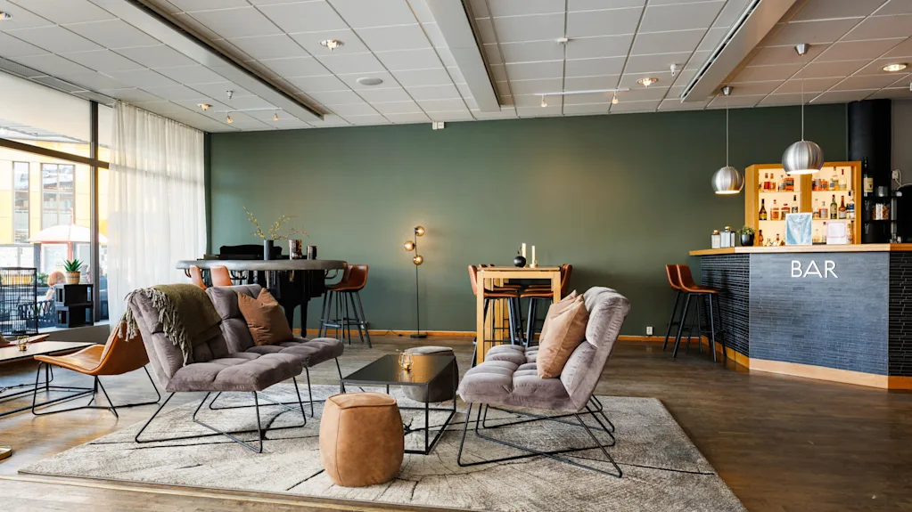 Kodikkaat lounge-tuolit lepäävät maton päällä pienen pöydän vieressä, lämpimästi valaistussa, modernissa baarissa. "BAR" näkyy selvästi tiskillä, jota ympäröivät juomahyllyt.