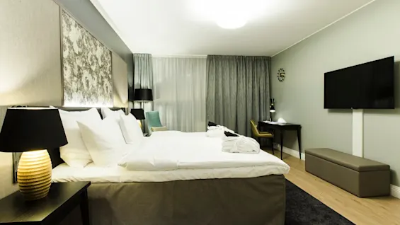 A neatly arranged hotel room with a large bed beside bedside lamps, facing a wall-mounted TV. A desk and chair are positioned near the window with draped curtains.