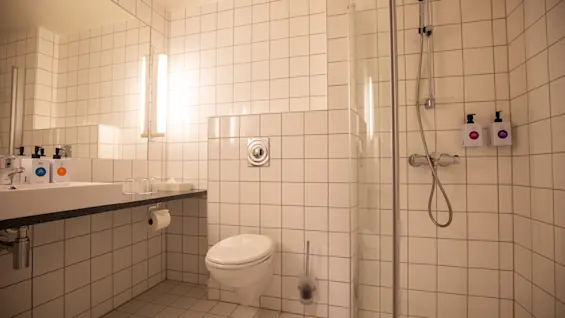 Et flislagt bad med vegghengt toalett, benkskap og speil. En dusj med glassdør og dispensere for såpe og sjampo er synlig ved siden av toalettet.