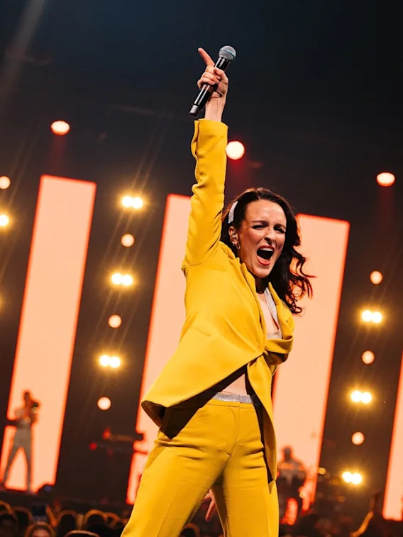 A female singer in a bright yellow suit holding a microphone up with high energy on stage under warm lighting.