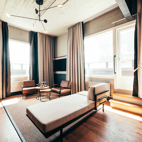 A modern living room features a chaise lounge, two brown chairs, and a round table with glasses. Neutral curtains frame large windows, and a contemporary ceiling light adds elegance.