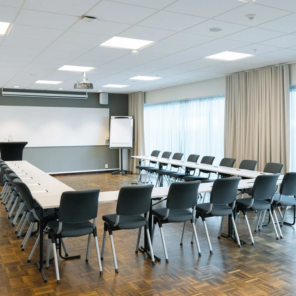 Konferensrum arrangerat i en U-form med stolar runt bord, en projektor i taket, en whiteboard på väggen och stora fönster med gardiner.