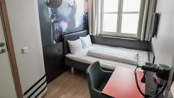 A small bedroom features a neatly made bed with white linens against a wall mural of a vintage microphone. A window provides natural light, and a desk with a hairdryer is nearby.