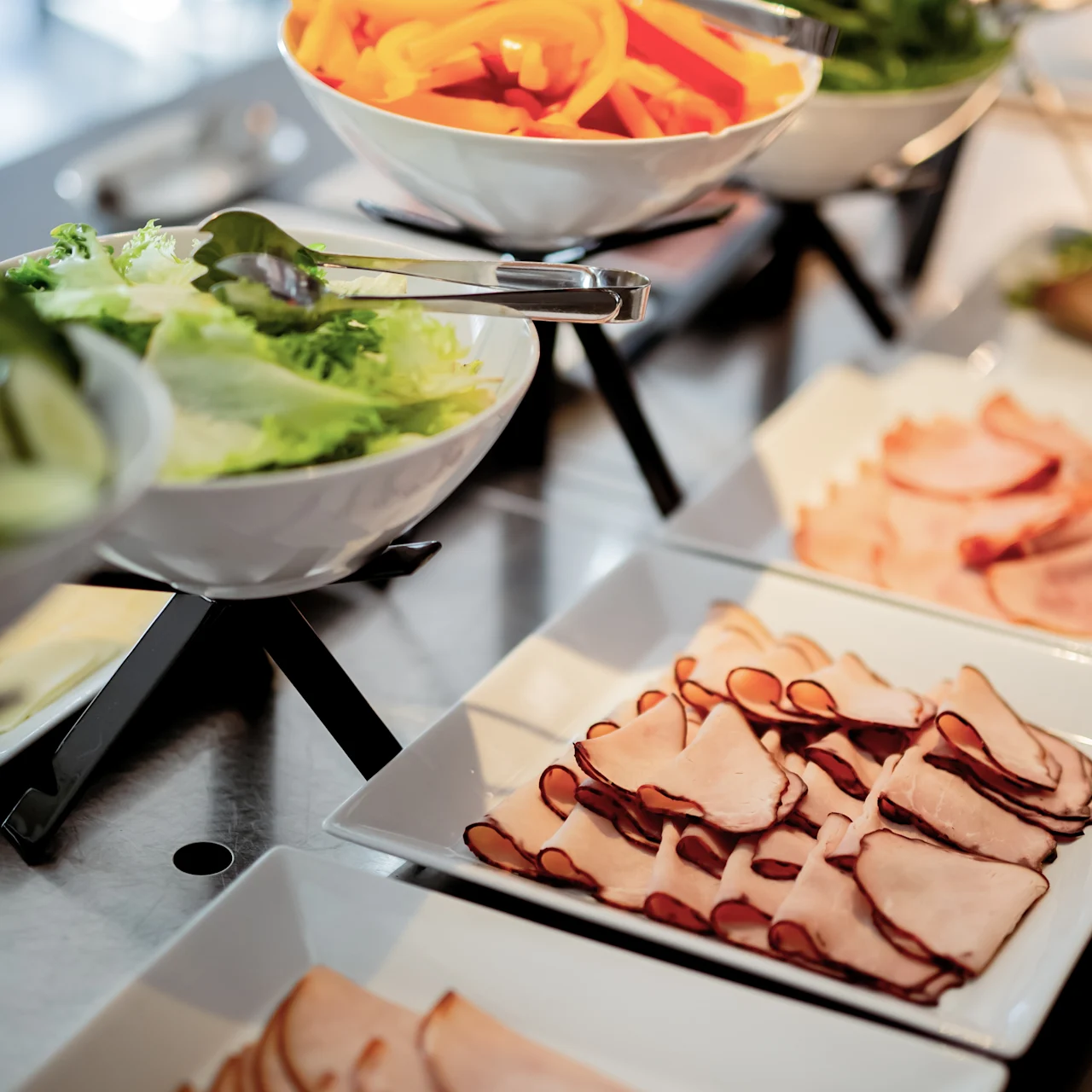Plates of sliced deli meats and bowls of fresh salad ingredients, including lettuce and bell peppers, are arranged on a buffet table with tongs for serving.