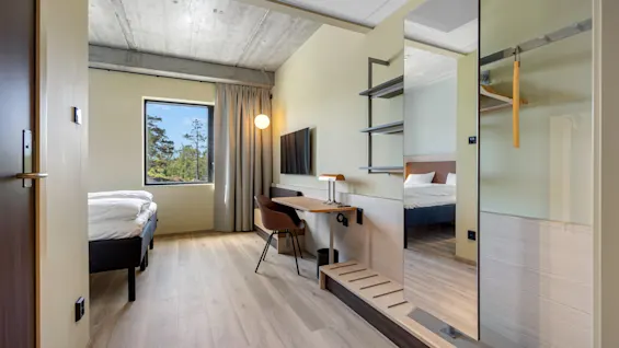 A modern hotel room with a bed, desk, TV, and a large mirror reflecting another bed and wardrobe.