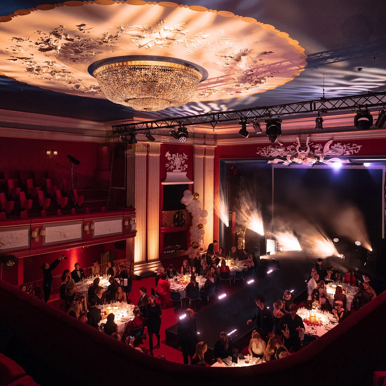 A chandelier hangs from a decorative ceiling over a dimly lit auditorium. People sit at round tables with candles, creating an elegant dining atmosphere on the main floor.