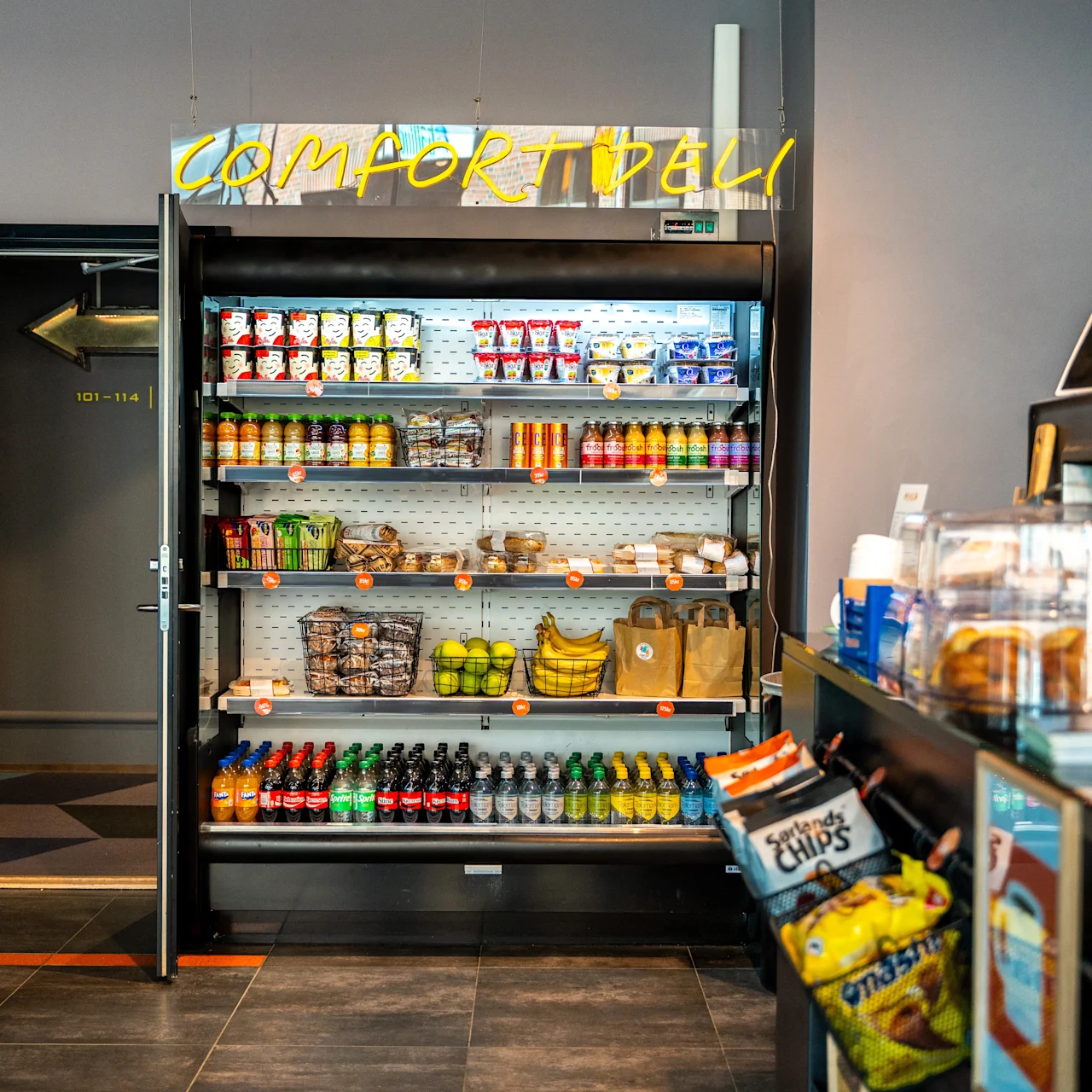 Jääkaappi esittelee välipaloja, juomia ja hedelmiä. Yläpuolella on neon-kyltti, joka lukee "COMFORT DELI". Lähellä on tiski, jossa sipsit ja leivonnaiset. Ovi merkinnällä "403-416" avautuu käytävään.