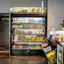 Jääkaappi esittelee välipaloja, juomia ja hedelmiä. Yläpuolella on neon-kyltti, joka lukee "COMFORT DELI". Lähellä on tiski, jossa sipsit ja leivonnaiset. Ovi merkinnällä "403-416" avautuu käytävään.