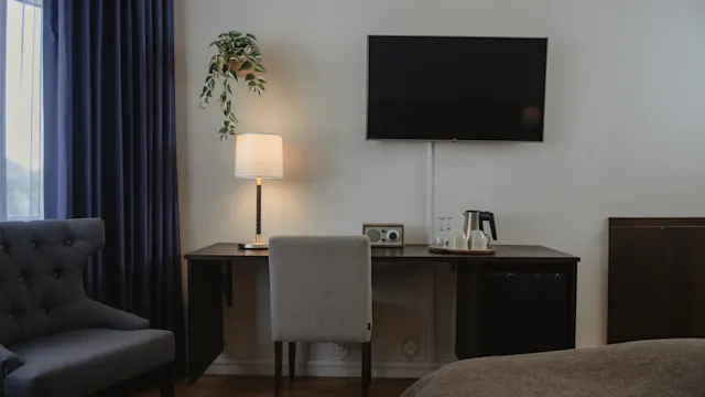 TV är monterad på en vit vägg, ovanför ett skrivbord med lampa, stol och växt. Vattenkokare och koppar finns i närheten. Mörka gardiner omger ett fönster.