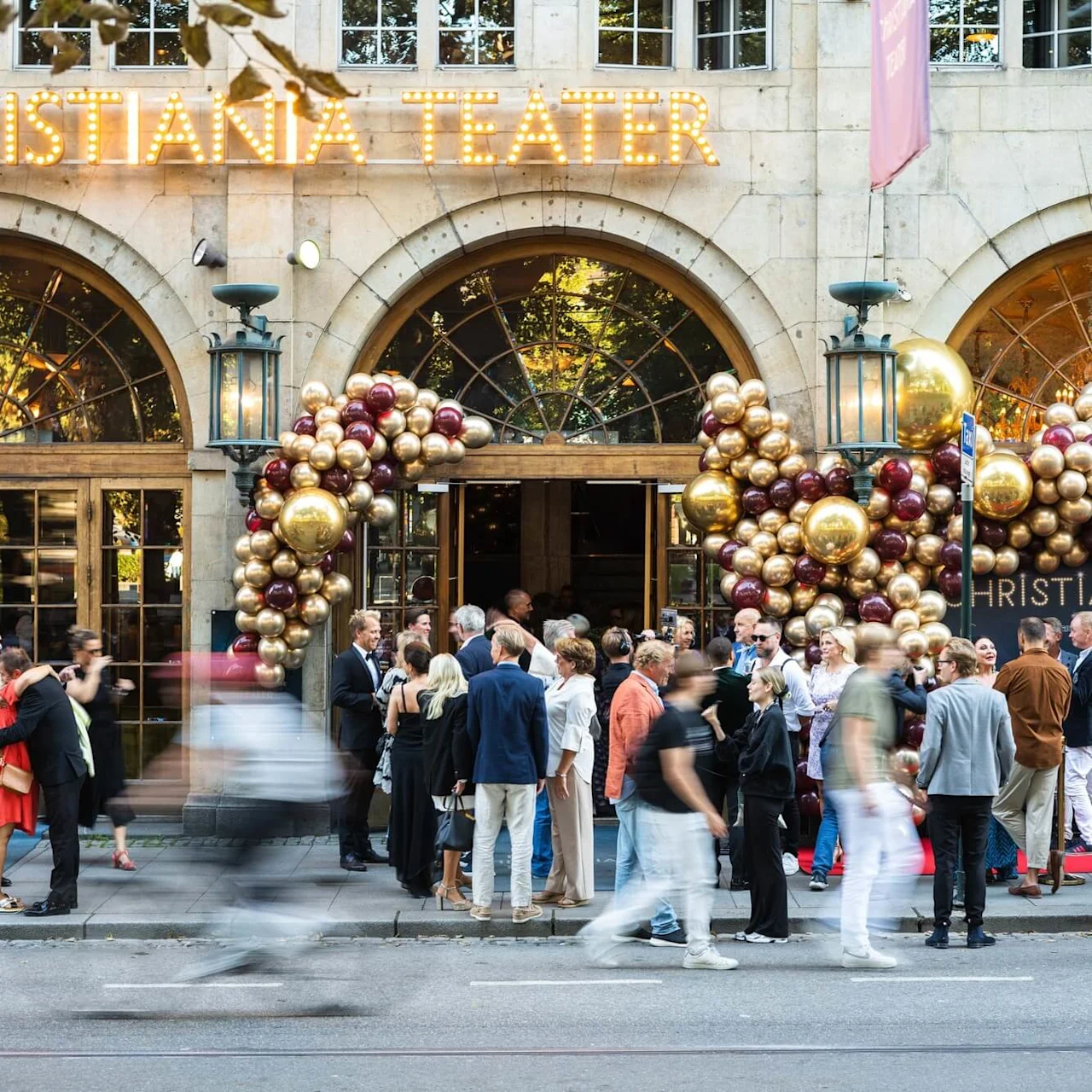 A crowd gathers outside a theater adorned with festive gold and burgundy balloons. "CHRISTIANIA TEATER" is prominently displayed in glowing lights above the entrance.