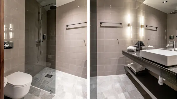 Moderne badeværelse med væghængt toilet og glasindelukket bruser til venstre; højre side har en vask på stentæller med håndklæder, lys og opbevaring nedenunder.