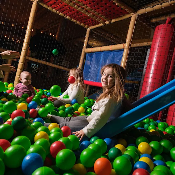 Barn som leker og smiler i et fargerikt innendørs ballbasseng ved en liten sklie.