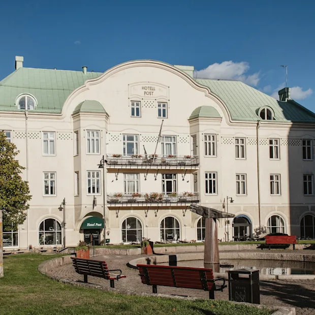 A grand white building with a green roof and balconies, labeled HOTELL POST and Hotel Post, stands behind a park with benches and a pond.