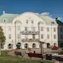 A grand white building with a green roof and balconies, labeled HOTELL POST and Hotel Post, stands behind a park with benches and a pond.
