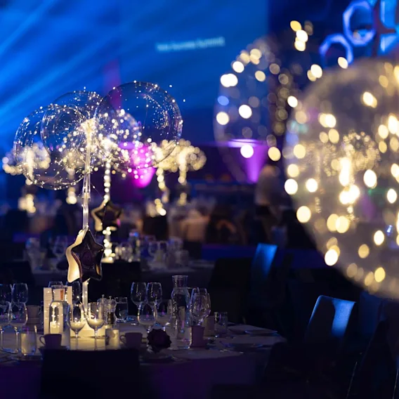 Bord är dukade för ett evenemang med elegant glas och glittrande ljusdekorationer. Blå och lila belysning skapar en festlig atmosfär. Text finns: "New | Secure Summit".