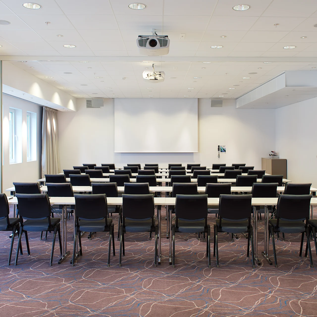 Rader med svarta stolar vända mot en stor vit projektskärm; rummet är väl upplyst med taklampor, och fönster till vänster släpper in naturligt ljus i en konferensrums miljö.