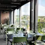 Et lyst restaurantområde med grønne stoler og mørke bord, med store vinduer som vender ut mot en by med hus og trær.