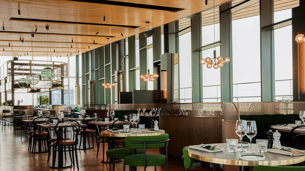 Modern restaurant setting; tables with green chairs and neatly set with glasses and cutlery. Large windows let in natural light, with pendant lights hanging from the ceiling.