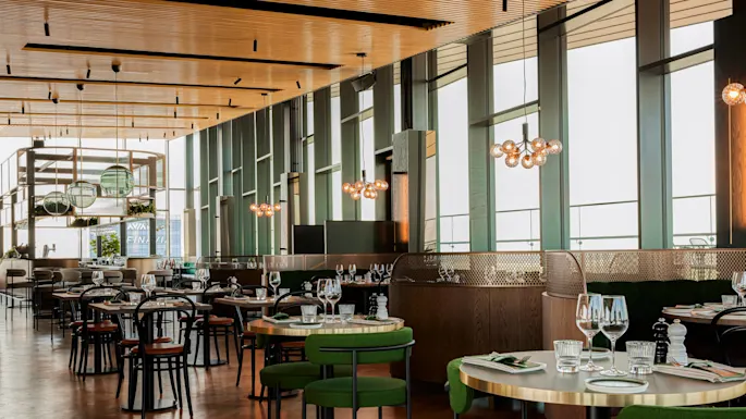 Moderne restaurantinnredning; bord med grønne stoler, pent dekket med glass og bestikk. Store vinduer slipper inn naturlig lys, med hengende taklamper.