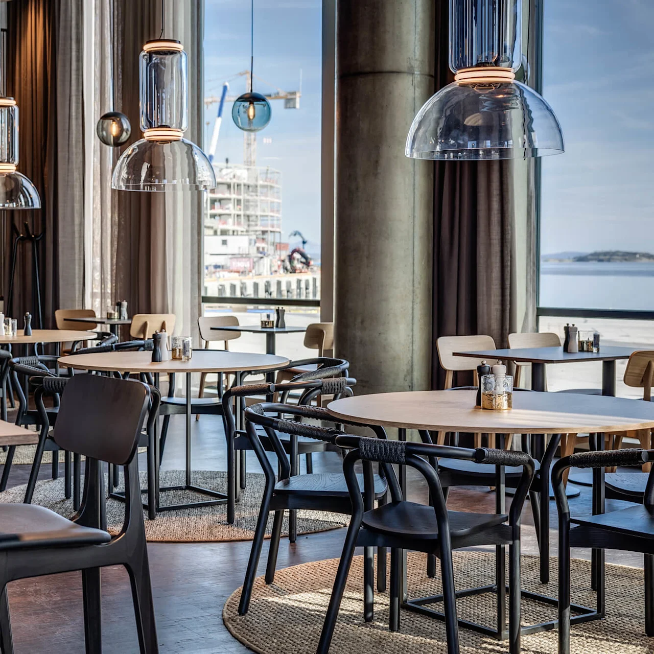Restaurant med moderne indretning: runde træborde, sorte stole, pendellamper hænger over. Store vinduer afslører en udsigt over konstruktion og vand, hvilket skaber en åben, luftig atmosfære.