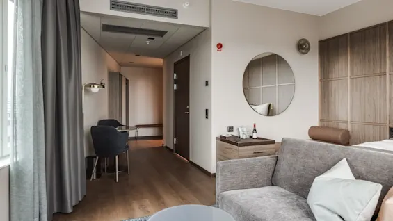 En grå sofa står i et moderne hotelværelse med trægulve, et rundt spejl og et lille bord med vin og glas under blød belysning.