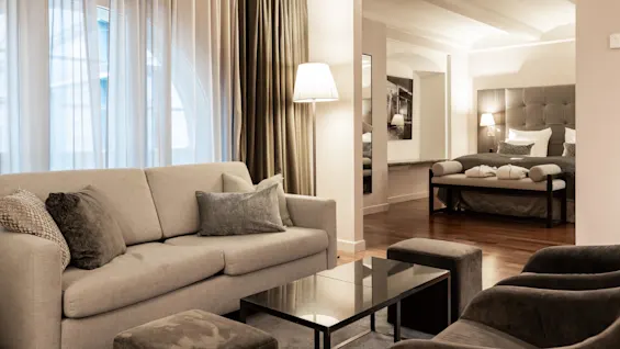 Beige sofa with pillows, positioned beside a glass coffee table; in the background, a bed with cushions and lamps, in a softly lit room with sheer curtains.