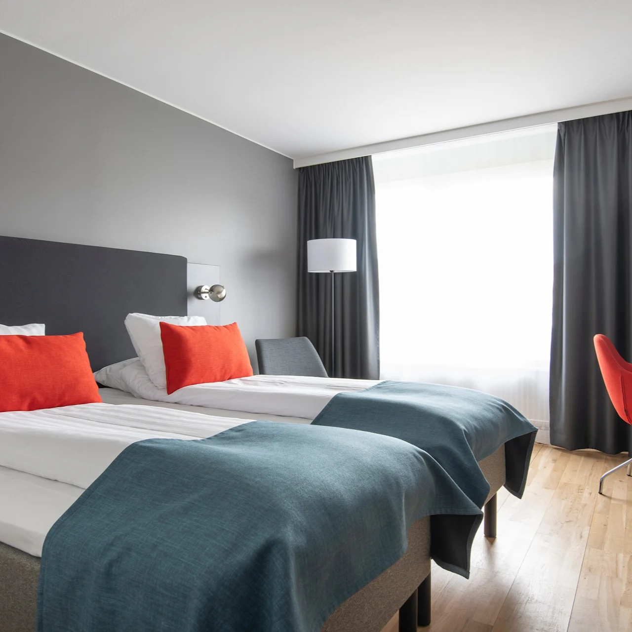 Två prydligt bäddade enkelsängar med röda kuddar och blå täcken i ett hotellrum. En röd stol och ett skrivbord står vid ett stort fönster med mörka gardiner.