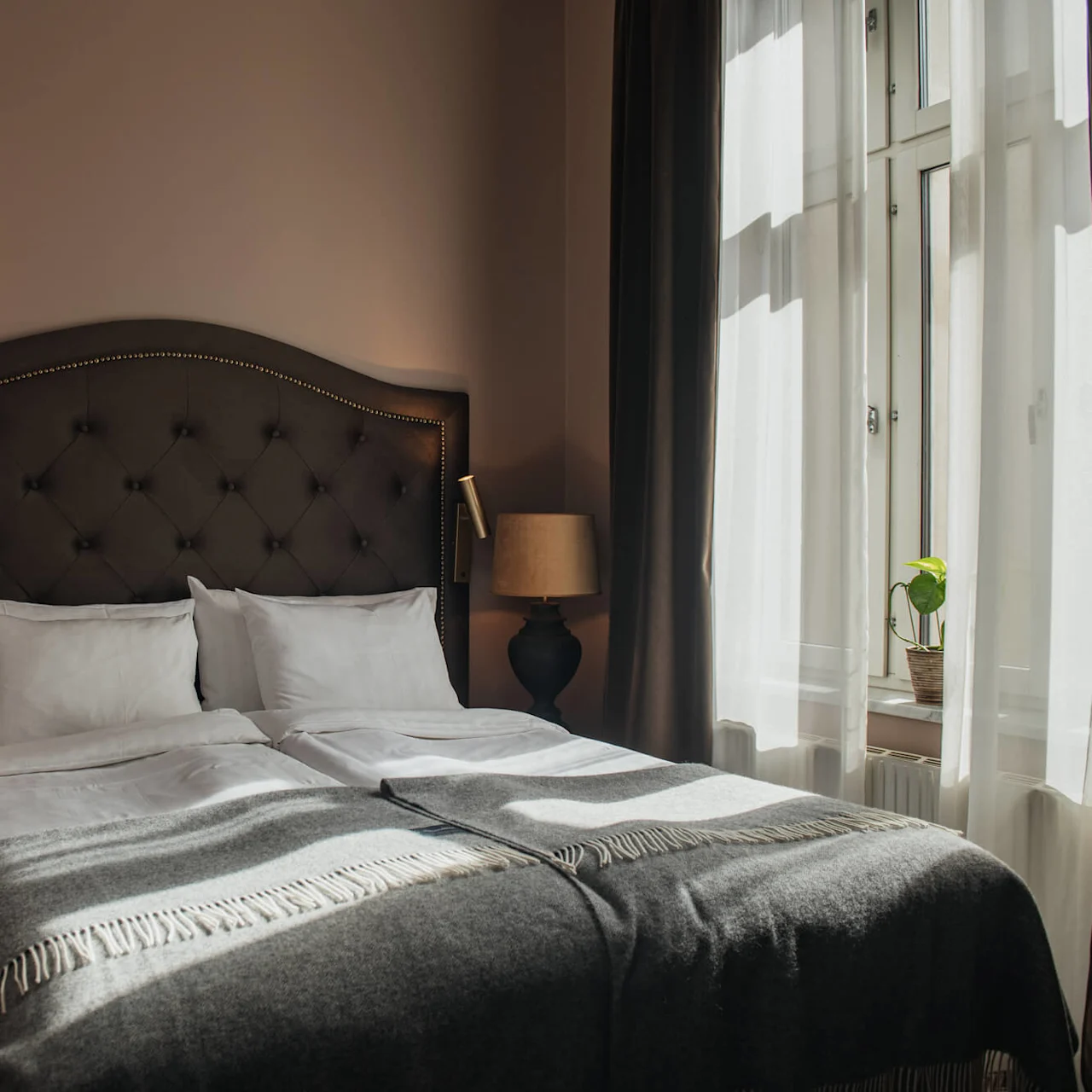 A neatly made bed with white pillows and a gray blanket is flanked by two lamps, situated in a warmly lit bedroom next to a large window with sheer curtains.