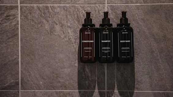 Tre mørke flasker merket balsam, sjampo og kroppsvask er montert på en teksturert grå flislagt vegg på et bad.