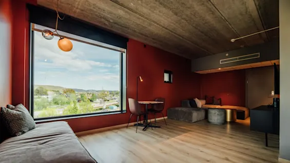 Et stort vindue giver udsigt til en naturskøn udsigt udendørs. Indenfor har et minimalistisk rum et bord med stole, en grå sofa og varme vægge. Trægulvet understreger den moderne indretning.