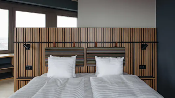 A bed sits neatly made, with white linens against a wooden slatted headboard, accented by patterned cushions and reading lights, in a modern, minimalistic bedroom with large windows.